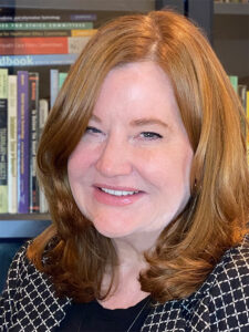 Head shot of Marin Gillis, a middle aged white woman with straight light to medium red hair sitting in front of a shelf of books