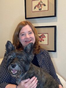 Marin Gillis sitting in a chair with small black dog on her lap, behind Marin is art depicting birds in the native american style of the pacific northwest